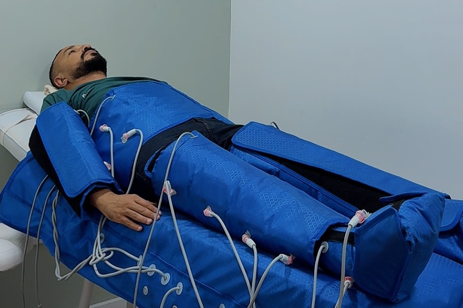 Patient allongé en cabine, jambes, bras et abdomen enveloppés dans les manchons bleus de pressothérapie reliés par leurs tubes